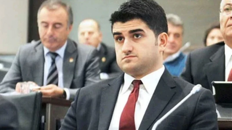 Young man in a dark suit and red tie sits attentively at a conference table, microphones in front, others blurred in the background.