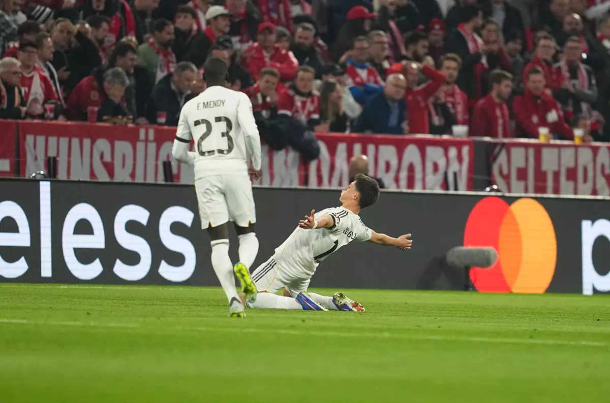 Soccer players in white kits on a grass pitch; one teammate stands with number 23, while another slides in a tackle as fans in red watches from the stands behind a digital ad board.
