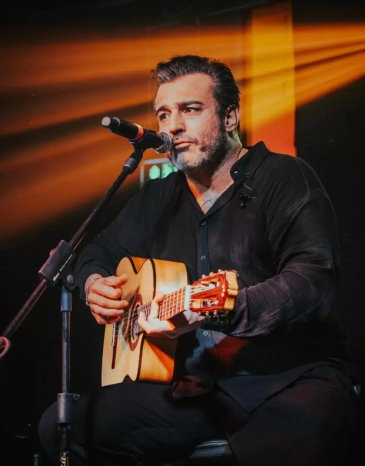 Male musician seated on stage, singing into a microphone while playing an acoustic guitar during a live performance