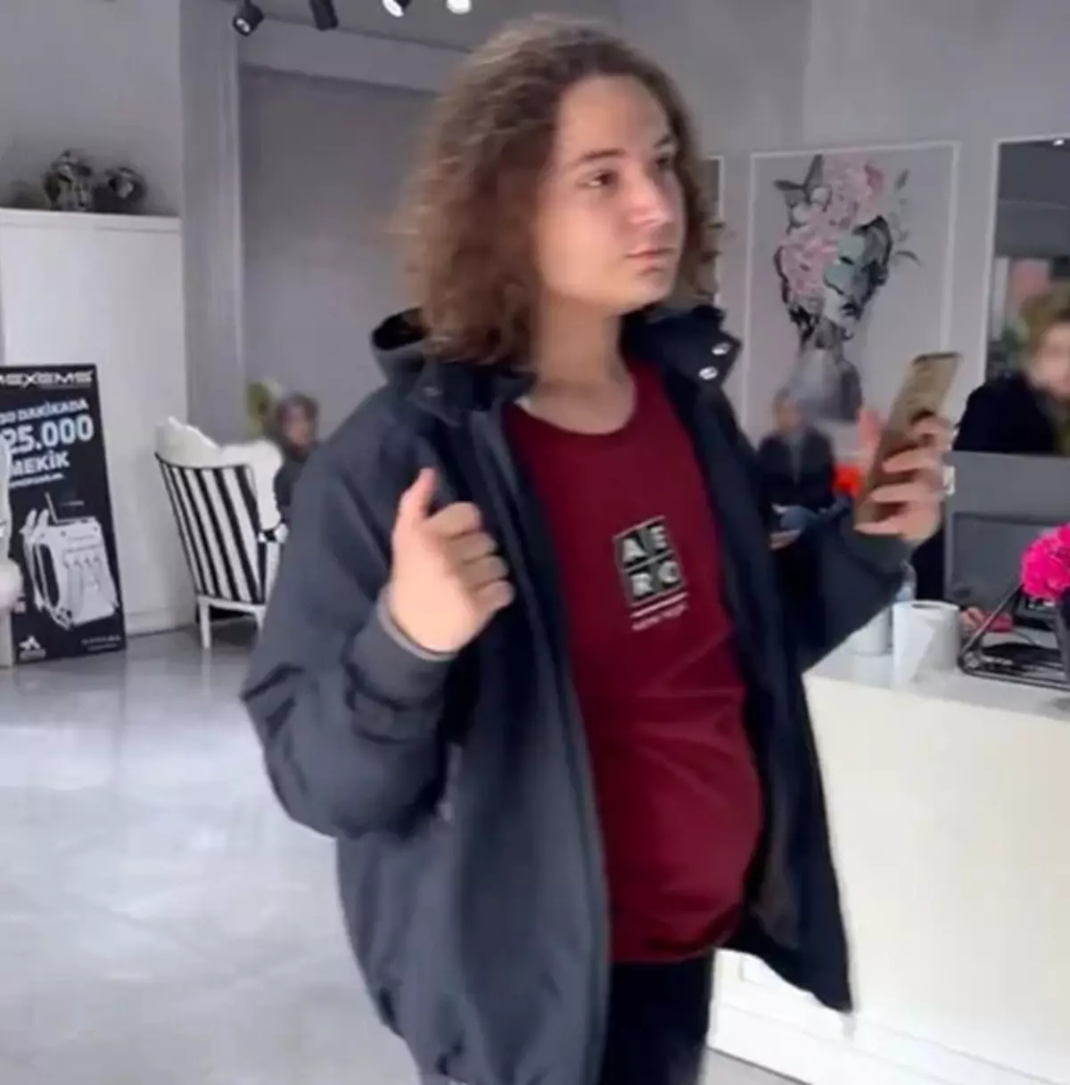 Young person with curly hair wearing a dark jacket and red shirt, holding a smartphone in a bright showroom.