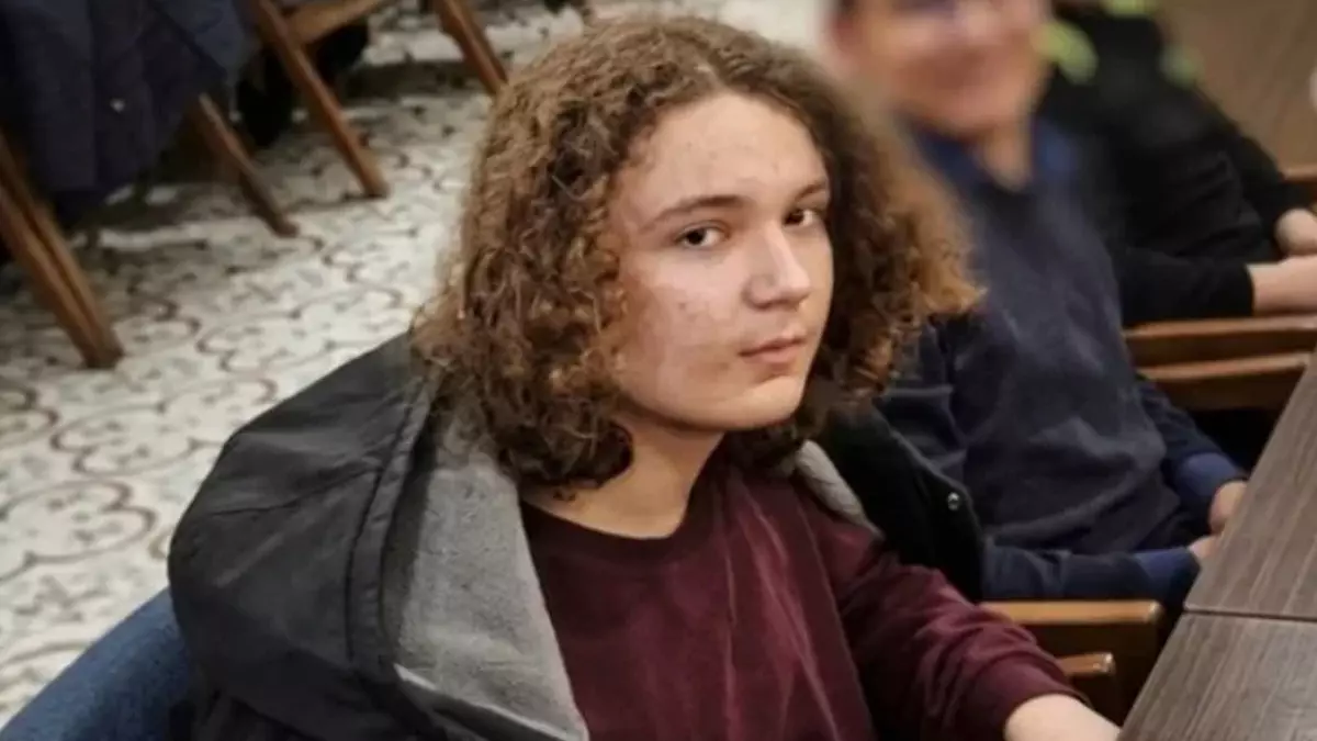 Young person with curly hair sits at a desk in a meeting or classroom setting, looking toward the camera.