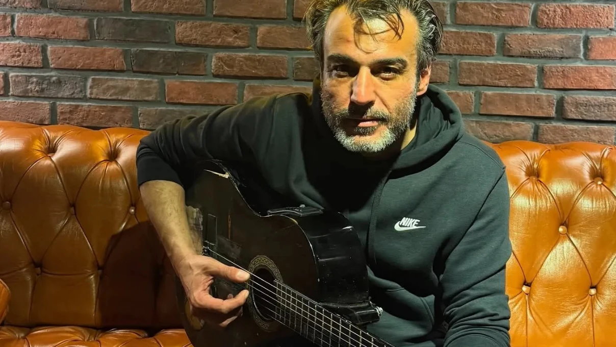 Man in a dark Nike hoodie sits on an orange tufted couch, playing a black acoustic guitar in front of a brick wall.