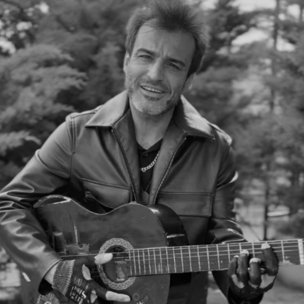 Smiling man in a leather jacket playing an acoustic guitar outdoors (black-and-white photo).