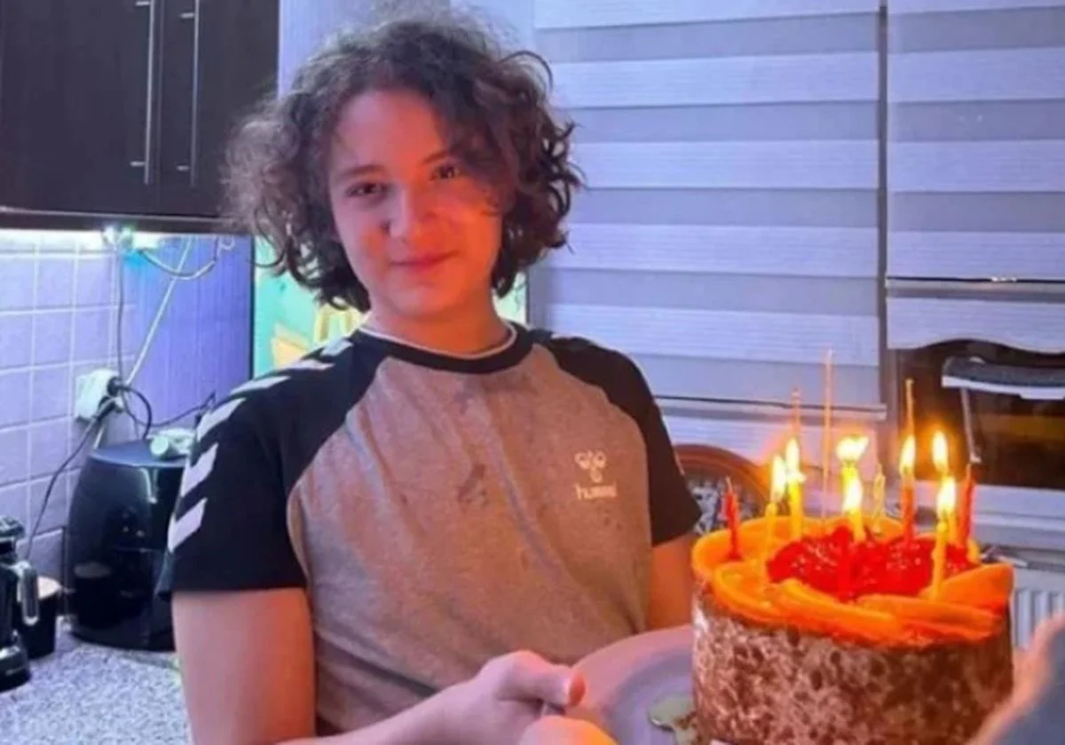 Child with curly hair in a kitchen holding a chocolate birthday cake with lit candles.