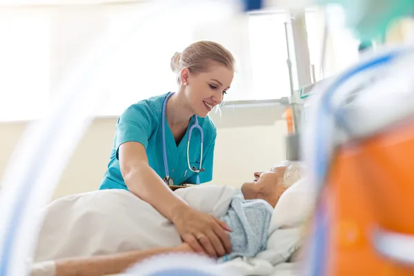 Nurse in teal scrubs with a stethoscope attends to a patient lying in a hospital bed.