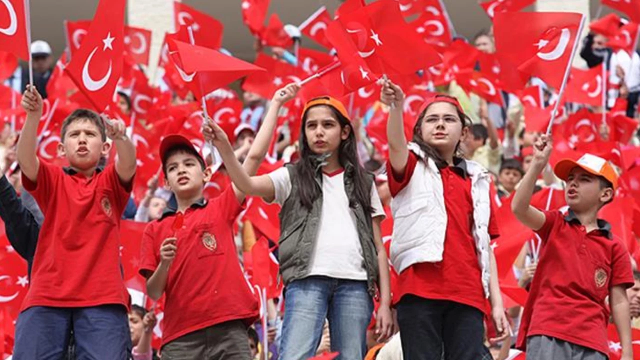 Group of children in red shirts waving Turkish flags at a crowded rally or parade.