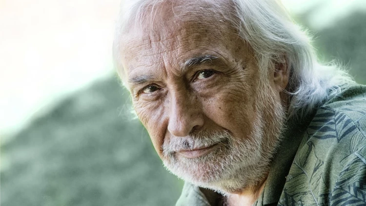 Close-up portrait of an elderly man with white hair and a white beard, outdoors with a soft green background.