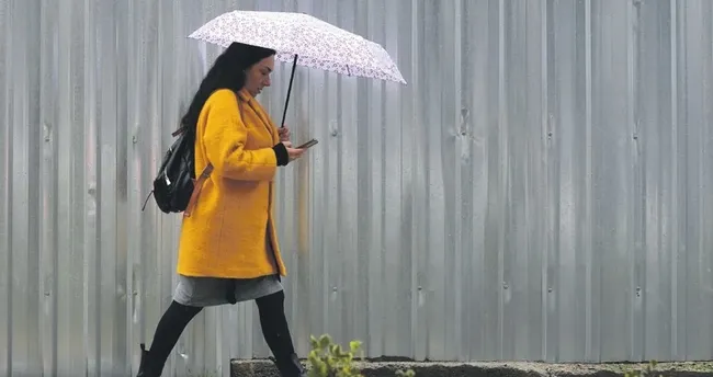 Woman in a bright yellow coat walks outdoors with a pink umbrella, glancing at her phone.