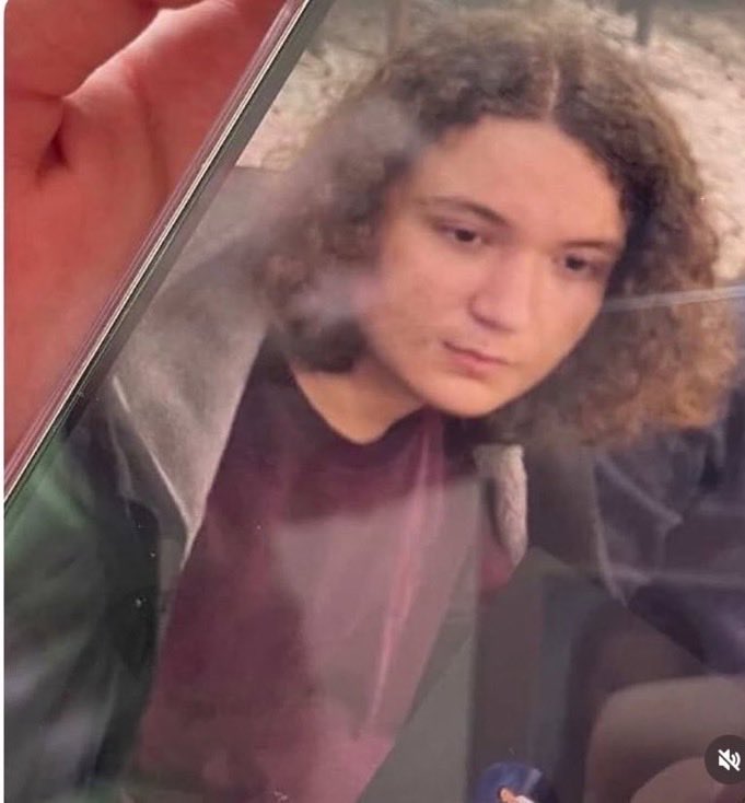 Person with curly hair looking down, seen through glass while holding a pink smartphone (reflection visible).
