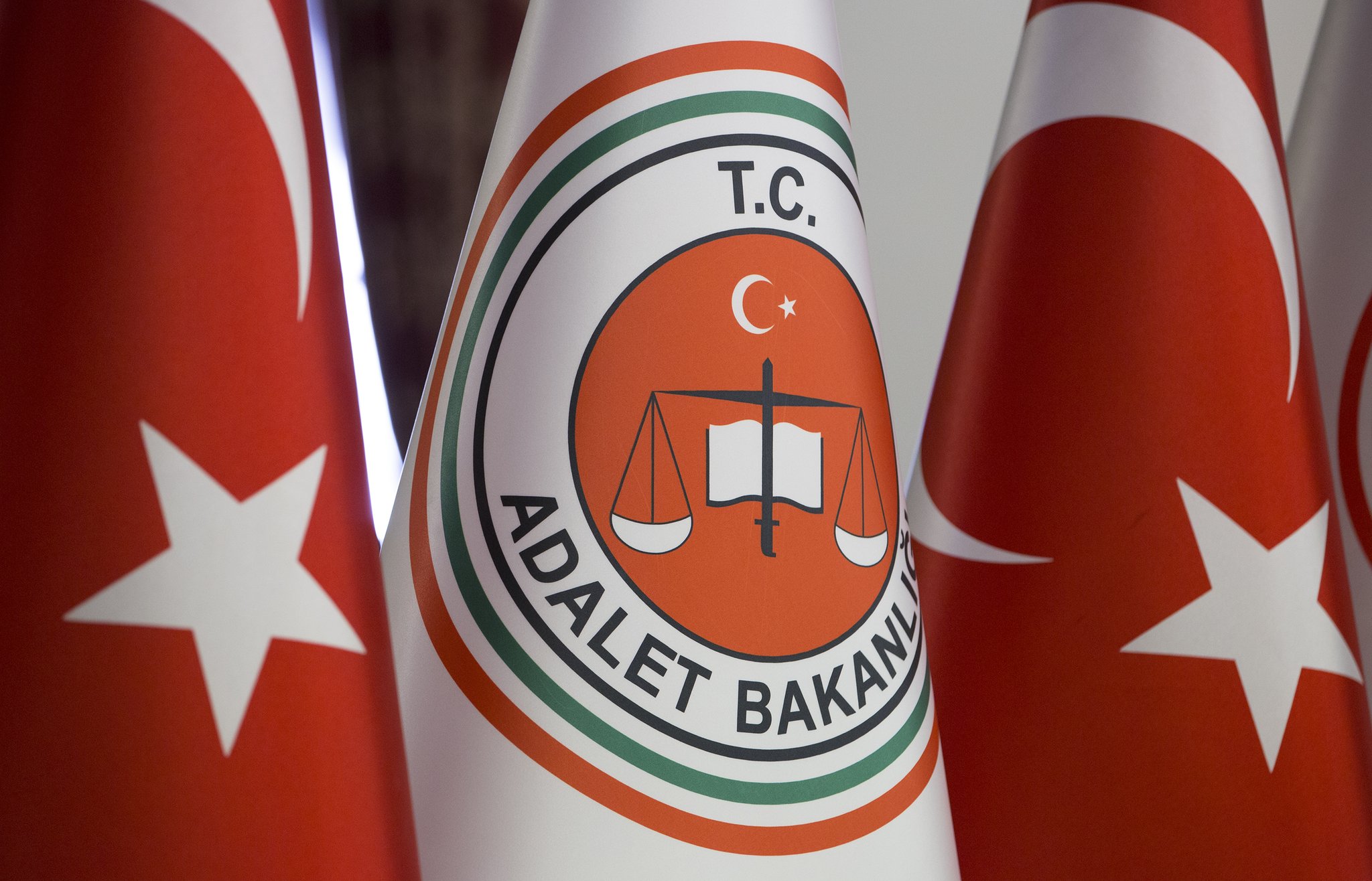 Close-up of the Turkish Ministry of Justice emblem (T.C. with scales of justice and book) on a white flag, flanked by red Turkish flags with white stars and crescents.