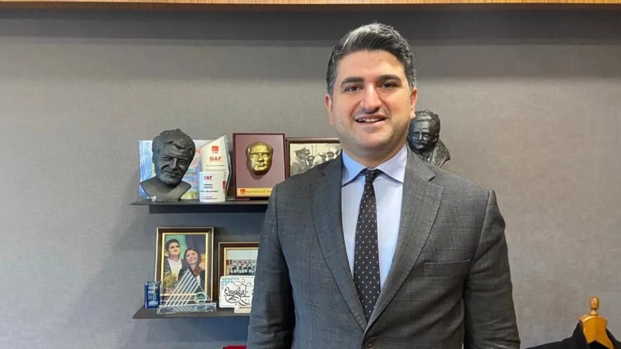 Man in a gray suit and blue dress shirt stands in an office with awards on a shelf behind him, smiling at the camera.