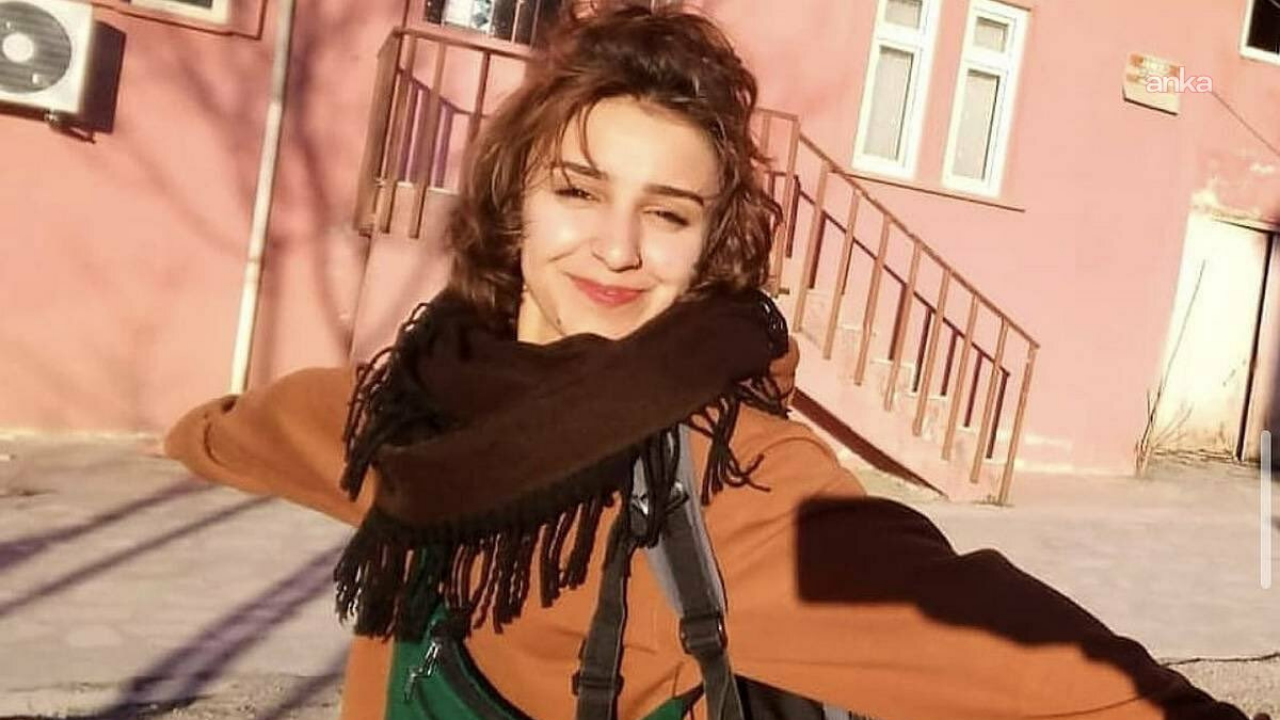 Smiling woman in a brown coat and scarf with arms outstretched in a sunny, pink building courtyard behind her.