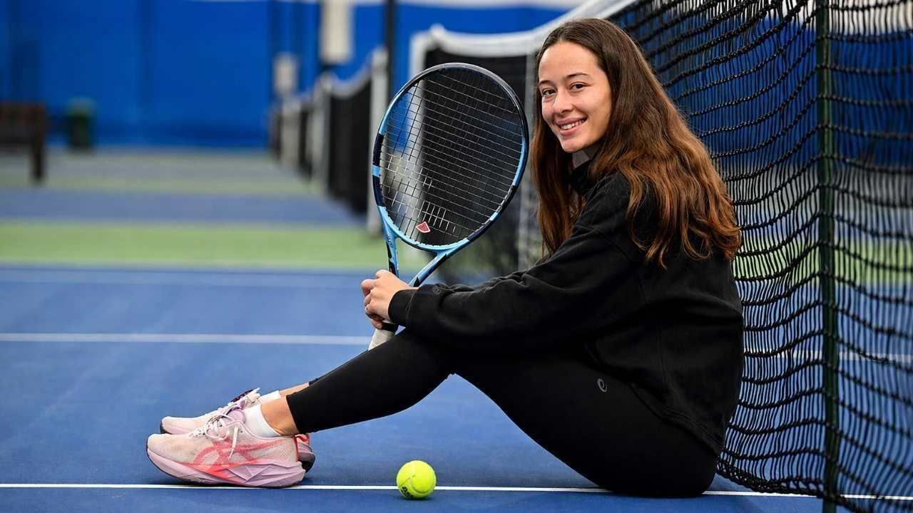 Woman sits on a blue tennis court, holding a racket beside a net with a tennis ball at her feet, smiling at the camera.