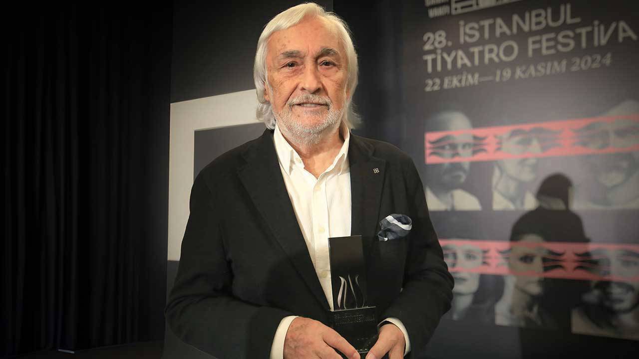 Older man in a black suit holding a trophy at a festival backdrop; looks toward camera with a subtle smile.