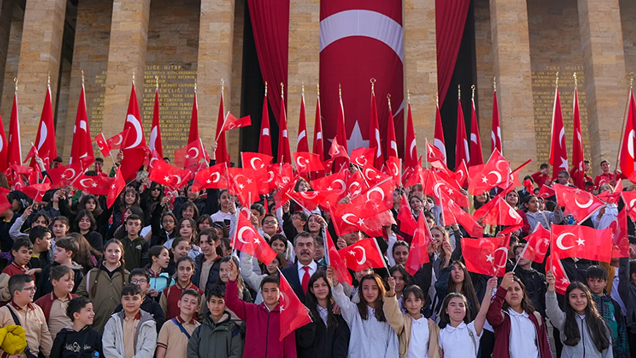 Crowd of students and adults waving Turkish flags at a patriotic ceremony in front of a building with a large central flag banner.