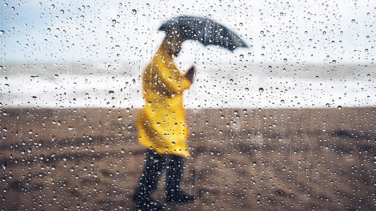 Person in a bright yellow raincoat walks on a rainy beach, holding a dark umbrella, seen through rain-speckled glass