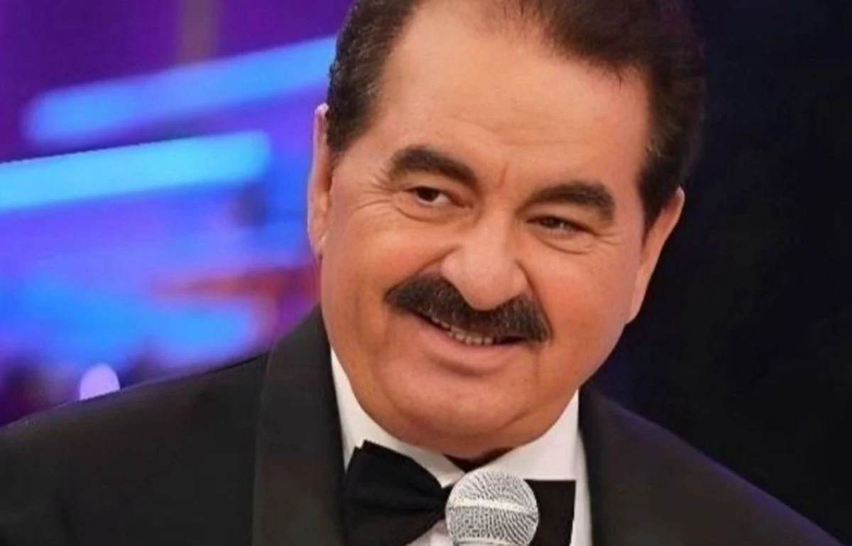 Smiling man in a tuxedo speaks into a microphone on stage with purple lighting in the background.