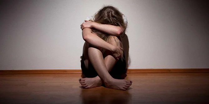 Person sitting on a wooden floor with knees drawn to chest and arms wrapped around them, head bowed in distress.