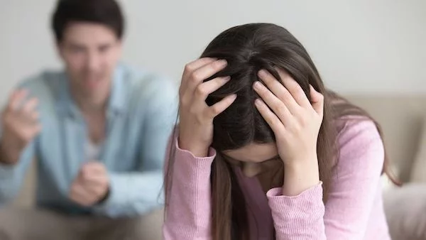 Young woman sits with her head in her hands, visibly distressed; a man in the background gestures as if arguing.