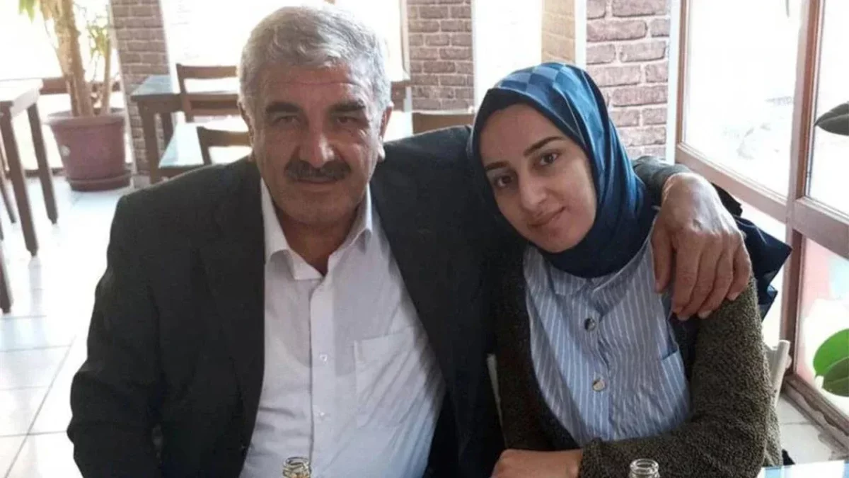 Two adults sit together at a cafe, the man in a suit with his arm around a young woman wearing a blue hijab and striped shirt.